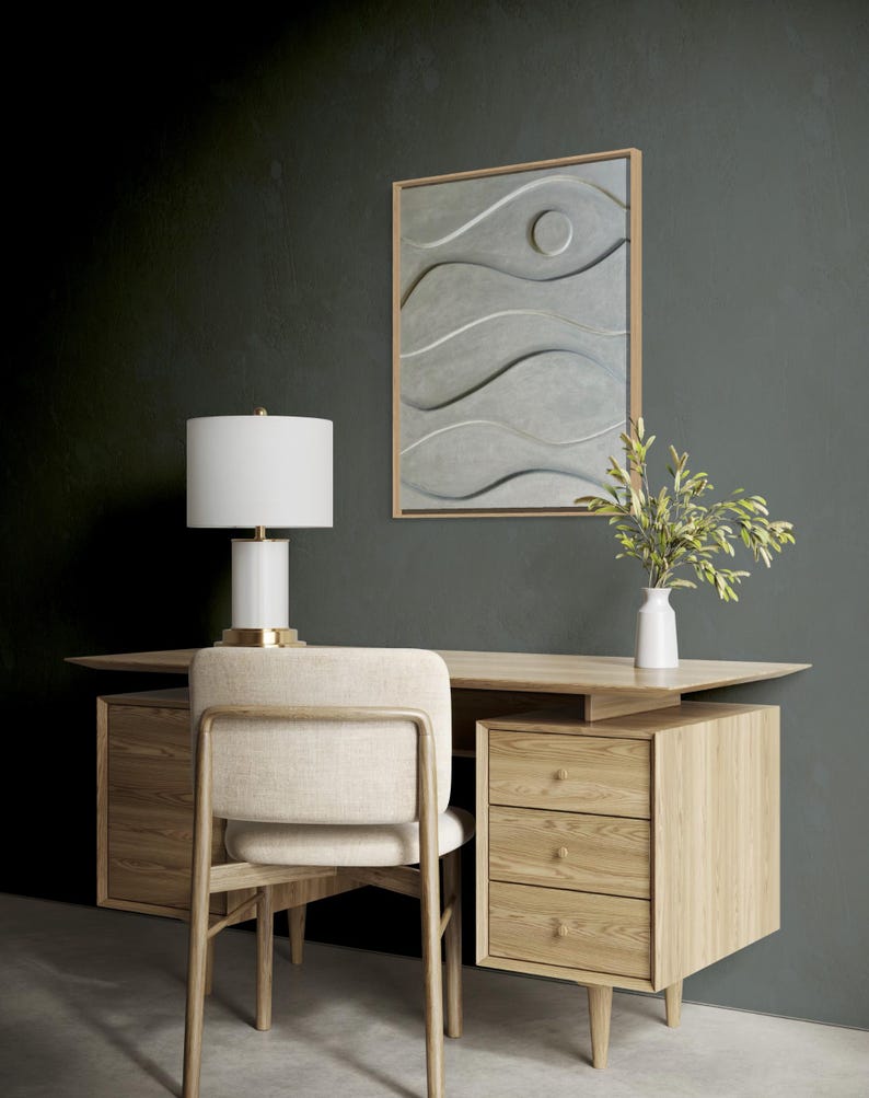 May include: A modern office setup featuring a light wood desk, chair, and a three-drawer cabinet. A white lamp with a gold base sits on the desk. A framed gray artwork with wave-like patterns hangs on the wall. A white vase holds green stems.