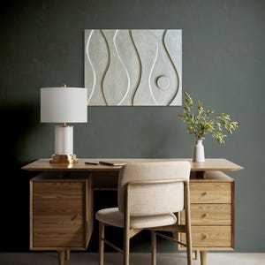 May include: A light wood desk with drawers and a matching chair. A white lamp with a gold base sits on the desk, along with a notebook and pen. A white vase with greenery and a decorative wall panel are also present.
