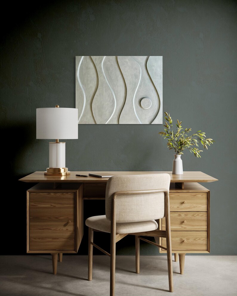 May include: A light wood desk with drawers and a matching chair. A white lamp with a gold base sits on the desk, along with a notebook and pen. A white vase with greenery and a decorative wall panel are also present.