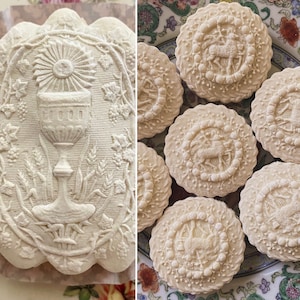 May include: A collection of intricately designed, white, embossed cookies. The cookies feature various religious symbols, including a chalice and a lamb. The cookies are arranged on a white plate with a floral pattern.