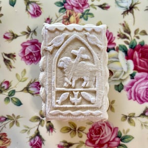 May include: A white, rectangular, embossed cookie with a design of a lamb, a cross, and two fish. The cookie is on a floral patterned surface.