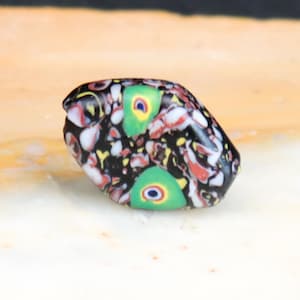 Può includere: Una perla di vetro colorata e decorata con motivi, con una base nera. La perla presenta accenti rossi, bianchi, gialli e verdi. Sono visibili due motivi verdi a forma di occhio. La perla è liscia e arrotondata.