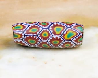 May include: A colorful glass bead with a geometric pattern of red, yellow, green, and blue. The bead is cylindrical and has a white background.