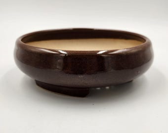 Op de afbeelding: Een ronde bonsai pot van bruin keramiek met een licht uitlopende rand. De pot is geglazuurd en heeft een drainagegat in de bodem.
