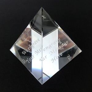 May include: A clear glass pyramid with the text "We engrave Your Message" and "Your Message Here" etched on the sides.
