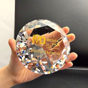 May include: A clear, faceted crystal award in a diamond shape. The award has gold lettering that reads "MD MEDICAL DATA CONSULTANTS INC" and "Doctor Thomson". The crystal reflects light, creating a sparkling effect.