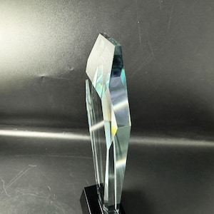 Op de afbeelding: Een heldere, gefacetteerde glazen trofee met een scherpe, hoekige bovenrand en meerdere geometrische oppervlakken die licht reflecteren. De transparante glasstructuur is stevig gemonteerd op een massieve, rechthoekige zwarte voet.