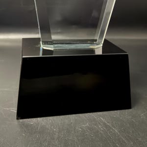 Op de afbeelding: Een trofee van helder glas met een gefacetteerd, taps toelopend bovendeel dat stevig op een massieve, rechthoekige zwarte voet staat. Het glazen onderdeel heeft scherpe, hoekige randen en een glad oppervlak, terwijl de voet ondoorzichtig zwart is.