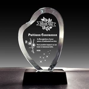 Puede incluir: Un premio de vidrio transparente en forma de corazón, montado sobre una base negra. El premio presenta el texto "THANK YOU" sobre el nombre "Pattiann Concannon" y un mensaje de agradecimiento. También se muestra el año "2025".