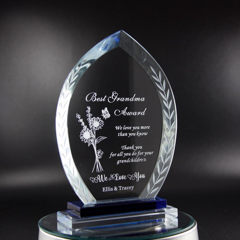 K&ouml;nnte beinhalten: Eine klare Glas-Auszeichnung mit blauer Basis, graviert mit dem Text "Best Grandma Award". Die Auszeichnung zeigt ein Blumenmuster und den Text "We love you more than you know. Thank you for all you do for your grandchildren. We love you. Ellis & Tracey".