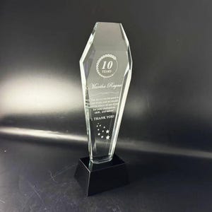 Op de afbeelding: Een heldere, gefacetteerde glazen trofee met een zwarte voet. De trofee is gegraveerd met de tekst 10 YEARS, Martha Ragna en een dankbetuiging, samen met een kleine stergrafiek onderaan, rechtopstaand tegen een donkere achtergrond.