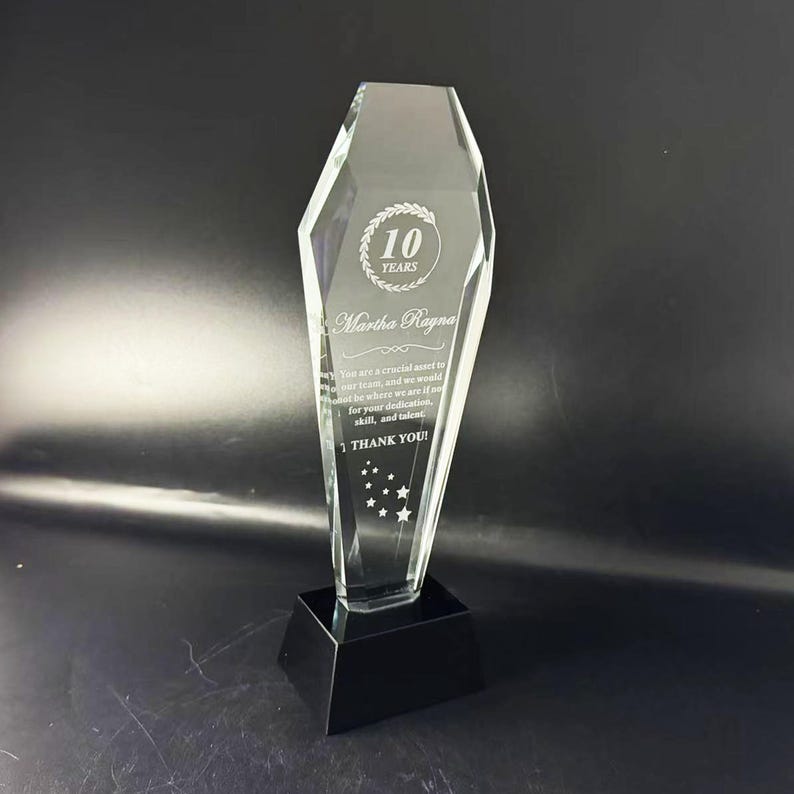 Op de afbeelding: Een heldere, gefacetteerde glazen trofee met een zwarte voet. De trofee is gegraveerd met de tekst 10 YEARS, Martha Ragna en een dankbetuiging, samen met een kleine stergrafiek onderaan, rechtopstaand tegen een donkere achtergrond.