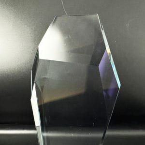 Op de afbeelding: Een helder, gefacetteerd glazen object of award met een veelzijdige, geometrische vorm. Het stuk heeft scherpe, hoekige randen en gladde, transparante oppervlakken die het licht reflecteren, waardoor duidelijke accenten en schaduwen ontstaan.