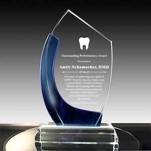 May include: A clear glass award with a blue accent, featuring the text "Outstanding Performance Award" and a tooth graphic. The award is presented to Amily Schamacher, DMD, and includes an inscription. The award sits on a clear, multi-faceted base.
