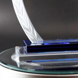 K&ouml;nnte beinhalten: Eine klare Glas-Troph&auml;e mit einem blauen Sockel und einem dekorativen, ge&auml;tzten Design. Die Troph&auml;e steht auf einem runden Glassockel.