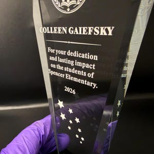 Op de afbeelding: Een heldere, wigvormige glazen trofee met de gegraveerde tekst: COLLEEN GAIEFSKY, For your dedication and lasting impact on the students of Spencer Elementary, 2026. De trofee is aan de onderkant versierd met kleine ge&euml;tste sterren.