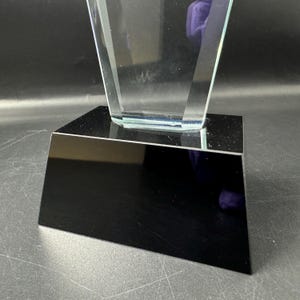 Op de afbeelding: Een heldere, transparante glazen trofee met een taps toelopend, geometrisch ontwerp. De glazen structuur is stevig gemonteerd op een massieve, rechthoekige zwarte voet met een gladde, reflecterende afwerking, wat zorgt voor een scherp contrast.