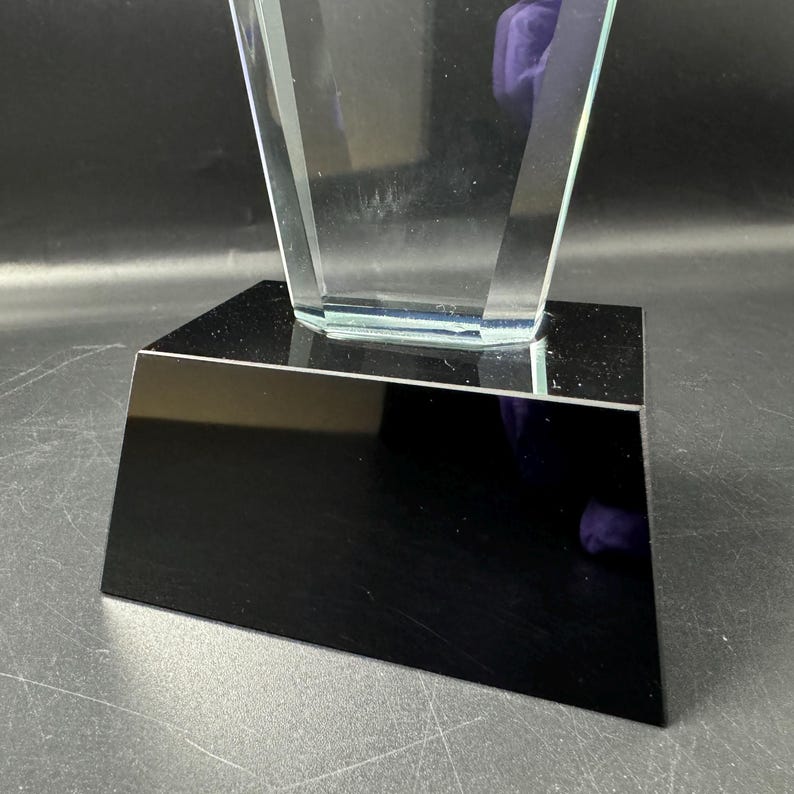 Op de afbeelding: Een heldere, transparante glazen trofee met een taps toelopend, geometrisch ontwerp. De glazen structuur is stevig gemonteerd op een massieve, rechthoekige zwarte voet met een gladde, reflecterende afwerking, wat zorgt voor een scherp contrast.