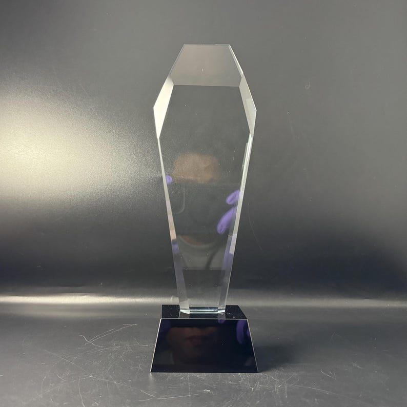 Op de afbeelding: Een heldere, transparante acryl award trofee met een hoog, veelzijdig, zeshoekig bovendeel dat taps toeloopt naar de top. De trofee is gemonteerd op een massieve, rechthoekige zwarte voet die een stabiele basis vormt voor het object.