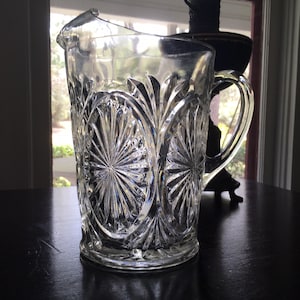 May include: A clear glass pitcher with a cut glass design featuring a repeating pattern of sunbursts. The pitcher has a handle and a wide mouth.
