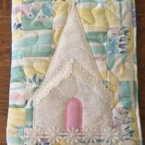 May include: A rectangular quilted fabric cover featuring a pastel patchwork pattern of yellow, blue, and white. Centered is an appliqued white fabric house shape with a pink arched door, white lace trim along the roofline, and a row of white daisy lace.