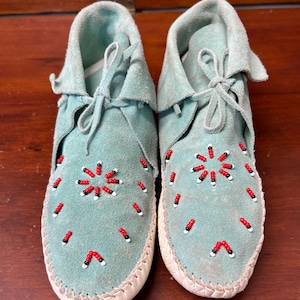 May include: A pair of light blue suede moccasins featuring a decorative pattern of small red, black, and white beads on the top. The footwear includes a tied lace closure at the front and a thick, light-colored stitched sole around the edges.