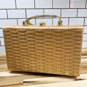 May include: A vintage rectangular handbag with a woven pattern in a light tan color. It features a gold-colored handle and clasp. The bag is sitting on a wooden surface.