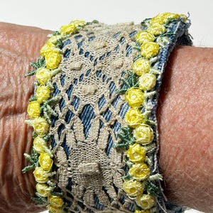 May include: A denim cuff bracelet with a beige lace overlay. The edges are adorned with yellow embroidered roses and green leaves. The bracelet is a unique handmade accessory.