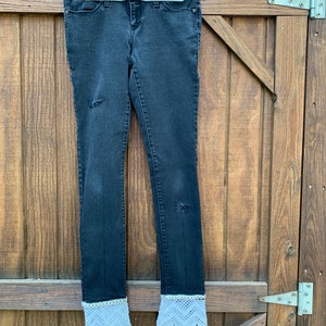 May include: Dark gray denim jeans with distressed details and white crochet lace cuffs. The jeans are hanging on a white hanger against a wooden background. The jeans have a classic five-pocket design.
