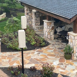 May include: Outdoor patio with a stone pathway and a covered seating area. The pathway is made of irregularly shaped stones in shades of brown and tan. Two beige covers are on black poles. The roof is dark gray.