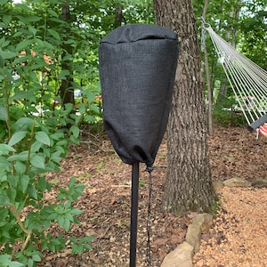 May include: A black outdoor speaker cover made of a textured fabric, secured with a drawstring. The cover is mounted on a black metal pole, set against a backdrop of lush green foliage and a hammock.