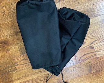 May include: Two black, cylindrical covers with drawstring closures. The covers are made of a smooth, opaque material. They are lying on a wooden floor with a natural wood grain pattern.