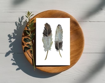May include: Two watercolor feather prints on a white card, displayed on a wooden plate. The feathers are in shades of brown, gray, and white. A sprig of green leaves and its shadow add a natural touch to the composition.