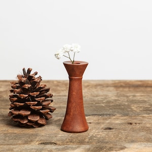 May include: A small wooden vase with a white flower inside, sitting on a rustic wooden surface next to a brown pine cone.
