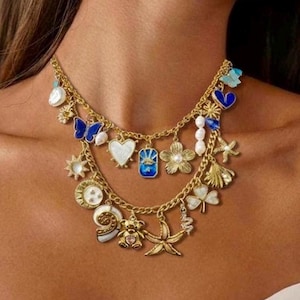 Puede incluir: Un collar de dijes dorado con dos hilos. Los dijes incluyen mariposas, corazones, flores y otras formas en azul, blanco y dorado. El collar se lleva alrededor del cuello.