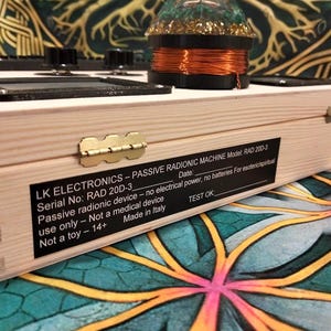 May include: A light-colored wooden box with a brass hinge and a black label that reads "LK ELECTRONICS - PASSIVE RADIONIC MACHINE." The box has knobs and a coil on top. The text on the label includes "RAD 20D-3" and "Made in Italy."