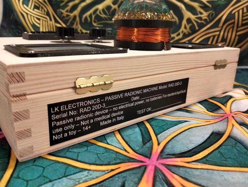 May include: A light-colored wooden box with a brass hinge and a black label that reads "LK ELECTRONICS - PASSIVE RADIONIC MACHINE." The box has knobs and a coil on top. The text on the label includes "RAD 20D-3" and "Made in Italy."