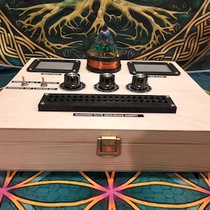 May include: A wooden box with a complex control panel featuring knobs, switches, and digital displays. A glass dome with a decorative element sits atop the box. The text "RADIONIC RATE SEQUENCE INSERT" is visible.
