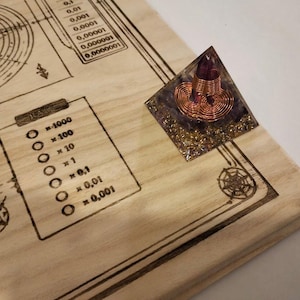 May include: A wooden surface with etched diagrams and numerical scales. A small, pyramid-shaped object with a purple crystal and copper wire coil sits on the surface. The wood has a light, natural color, and the etched lines are dark brown.