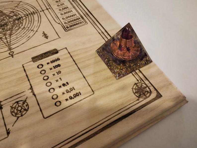 May include: A wooden surface with etched diagrams and numerical scales. A small, pyramid-shaped object with a purple crystal and copper wire coil sits on the surface. The wood has a light, natural color, and the etched lines are dark brown.
