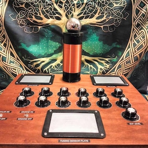 May include: A wooden device with multiple knobs, switches, and a central cylindrical component wrapped in copper wire. A silver sphere sits atop the cylinder. The device has a "Tuning Sensor Plate" label and is set against a tapestry with a tree design.