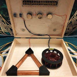 May include: Open wooden box with electronic components. Inside, a copper triangle, a coil of wire, and various circuits are visible. The box is open, revealing the internal wiring and components.