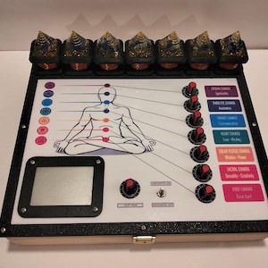May include: A rectangular device with a white control panel featuring a line drawing of a person in a seated pose. Seven colored chakra circles align with the body, connected to seven black knobs with red accents. Seven pyramid-shaped orgonite pieces sit along the top edge.