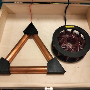 May include: A wooden box contains a copper triangle with black corner supports and a circular coil of red wire. A black wire and a red wire extend from the components. A yellow sticker on the coil reads "LK 23.03.19".