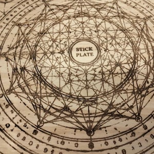 May include: A wooden plate with a complex geometric design etched into its surface. The design features interconnected lines, circles, and a central text that reads "STICK PLATE". The plate has a light brown color.