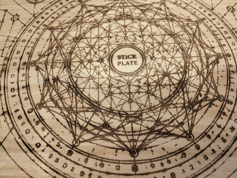 May include: A wooden plate with a complex geometric design etched into its surface. The design features interconnected lines, circles, and a central text that reads "STICK PLATE". The plate has a light brown color.