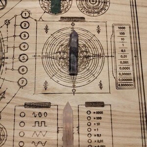 May include: A wooden board with etched geometric designs and text, including "UNIVERSAL SYSTEM INTERFACE" and numerical scales. Three crystal points in green, purple, and pink are arranged on the board, suggesting a spiritual or metaphysical theme.