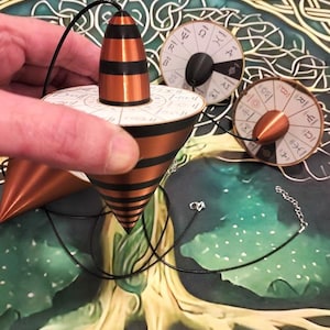 May include: A conical copper and black striped pendulum with a white circular top featuring Greek letters and symbols. The pendulum is attached to a thin black cord. Two additional similar pendulums with white circular tops are visible nearby.