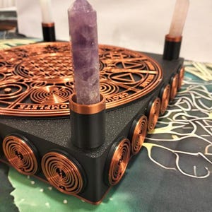 May include: A black square base featuring a copper-colored circular geometric design on top and copper spiral accents on the sides. Three upright crystal points, including a central purple amethyst, are held in black and copper cylindrical mounts.