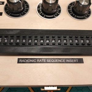 May include: Close-up of a wooden panel with black dials, switches, and a black digital display. The display shows a series of numbers and letters. The text "RADIONIC RATE SEQUENCE INSERT" is printed below the display.